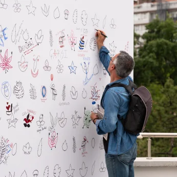 Bir alanın duvarında, çeşitli desenler ve yazılarla dolu bir yüzey yer almakta. Duvarın üst kısmında farklı bitki desenleri, yapraklar ve soyut şekiller var. Bazı çizimlerde kalp, gül, ve göz gibi semboller de dikkat çekiyor. Ortada canlı renklerle yazılmış kelimeler ve cümleler yer almakta. Bir birey, elinde bir şeyler tutarak, duvarın üst kısmına doğru eğilerek bir şeyler yazıyor. Bu kişi, mavi bir gömlek giymekte ve omzunda bir çanta var. Arka planda, yoğun bir yeşillik ve birkaç bina görünmekte. Bireyin etkinlikte aktif olarak yer aldığı, yaratıcı bir ortamda bulunduğu hissi uyandırıyor.