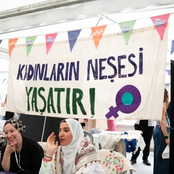 Bir etkinlik alanında, arka planda büyük bir bez banner asılı. Banner üzerinde koyu mavi ve yeşil renklerle yazılmış "Kadınların Neşesi Yaşatır!" ifadesi yer alıyor. Alt kısmında sembolik bir kadın işareti bulunuyor. Banner’ın altında, iki kişi oturuyor. Birinci kişi, gülümseyerek kameraya doğru bakıyor, siyah bir bluz giymiş ve başında desenli bir örtü var. İkinci kişi, kuş tüyü detayları olan bir baş örtüsü takıyor, elini ağzına götürmüş ve konuşuyor gibi görünüyor. Her iki kişi de yanlarında su şişeleri ve çeşitli materyallerle dolu bir masa var. Etkinlik alanında, arka planda başka katılımcılar ve stantlar dikkat çekiyor, çeşitli renklerde bayraklar asılı. Genel atmosfer canlı ve hareketli.