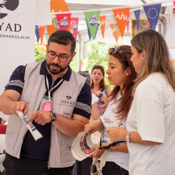 Etkinlik alanında bir grup insan yer almakta. Merkezde, bir kişi elinde bir broşür tutarak diğerlerine bilgi veriyor. Bu kişi gri bir yelek giyiyor ve kafasında gözlük var. Diğer iki kişi, dikkatle dinlemekte ve broşüre bakıyor. Birisi beyaz bir tişört giymekte, diğeri ise daha koyu renkli bir üst giysi tercih etmiş. Arka planda renkli bayraklar asılı durmakta ve çeşitli stantlar görünmekte. Alanın aydınlık olduğu, katılımcıların etkinlikten memnun olduğu hissedilmektedir.