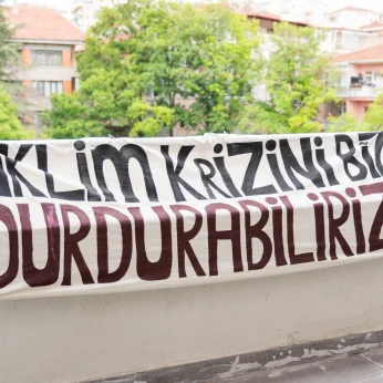 Bir verandalarda, bir ip üzerine asılmış geniş bir bez parçası bulunmaktadır. Bez parçasında dikkat çekici şekilde büyük harflerle yazılmış yazılar yer alıyor. Üst kısımda "İKLİM KRİZİNİ" ifadesi, alt kısımda ise "DURDURABİLİRİZ" ifadesi bulunuyor. Yazıların renkleri siyah ve derin bir kırmızı tonunda olup, zemin bezi açık renktedir. Arka planda, çeşitli ağaçlar ve binalar görünmektedir; bu binaların bazı pencerelerinde insanlar görülebilir. Genel olarak, dış mekan olduğu ve çevrede doğal bir atmosferin bulunduğu hissedilmektedir.