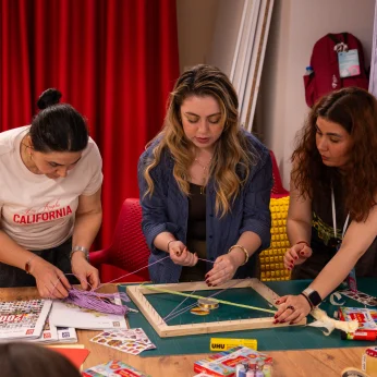 Bir grup insan, bir masa etrafında bir araya gelerek el sanatlarıyla uğraşmaktadır. Masanın üzerinde, çeşitli renklerde ipler ve malzemeler dikkatlice yerleştirilmiştir. Bir kişi, mavi gömlek giymekte olup, bir çerçeve şeklinde düzenlenmiş ipleri dikkatlice yerleştirirken konsantre bir şekilde çalışmaktadır. Diğer bir kişi, beyaz bir tişört giymekte ve o da ipleri yönlendirmekte. Yanındaki kişi ise daha koyu bir elbise içinde, masada serili olan malzemeleri düzenlemekte ve zaman zaman diğerlerinin çalışmalarına göz atmaktadır. Arka planda, kırmızı perdeler ile bir çanta ve çeşitli araçlar yer almakta, ortamda yaratıcılıkla dolu bir atmosfer oluşmaktadır. Herkes, el becerilerini kullanarak ortak bir projeye odaklanmış bir şekilde görünmektedir.