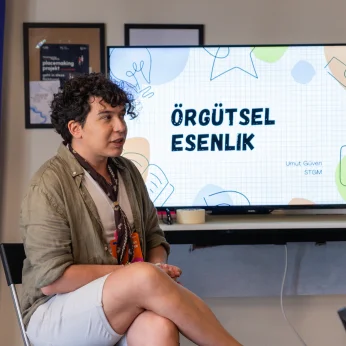 Bir konuşma sırasında bir kişinin otururken görünümü dikkat çekiyor. Kişi, açık renkli, kısa bir şort giymekte ve üzerine açık yeşil tonlarında bir gömlek giymiş. Üzerinde ince bir şal veya fular yer almakta. Yüzü hafifçe yana dönmüş ve konuşmaya dalmış durumda. Arkada bir ekran var; bu ekranın üzerinde "ÖRGÜTSEL ESENLİK" yazılı. Ayrıca, ekranın etrafında çeşitli grafikleri andıran çizim ve renk blokları gözlemleniyor. Arka planda, Avrupa Birliği bayrağı ve bazı çerçevelenmiş belgeler dikkat çekiyor, bu belgeler içerik açısından görünmüyor fakat organizasyon bağlamında bir ortam oluşturuyor. Kişinin oturduğu yerde bir masa ve sandalyeler mevcut, ortam rahat ve samimi bir havaya sahip.