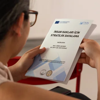 Bir kişi, bir masanın yanında oturmakta ve iki elle bir belgeyi kavramış durumda. Belgenin ön yüzünde "İNSAN HAKLARI İÇİN STRATEJİK DAVALAMA" yazıyor. Üst kısımda belgeleri hazırlayan kişilerin isimleri de belirtilmiş. Kişinin kıyafeti açık renkli, üst kısmı dantel veya dikiş detaylarına sahip. Arkada duvarda, sade bir ortam olduğu anlaşılıyor. Kişinin oturduğu sandalyenin rengi kırmızı ve belgenin yanında bir cep telefonu görünmekte. Kıymetli bir konunun ele alındığı belirgin bir atmosfer mevcut.