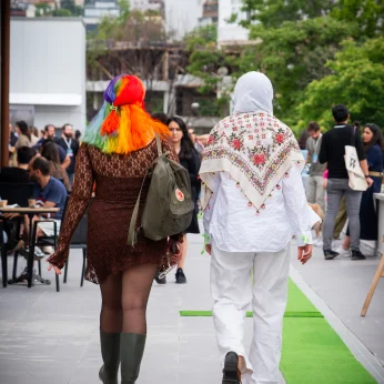 İki kişi, şehir ortamında yürüyüş yaparken arka planda yer alıyor. Birinci kişi, uzun, kahverengi, dantel detaylı bir kıyafet giymekte ve rengarenk, dalgalı bir saç stiline sahip. Sırtında yeşil bir sırt çantası var ve yeşil lastik çizmeler giymiş. İkinci kişi, beyaz bir elbise üzerine geleneksel desenli bir örtü sarmış ve başını örtmüş. Yürüyüş yolu, yanlarında yer alan yeşil bir halı parçası ile dikkat çekiyor. Arka planda, diğer insanlarla dolu bir ortamda kalabalık bir görüş alanı var; farklı kıyafetler giyen insanlar oturup hareket halindeler. Binalar ve yeşil ağaçlar, çevreyi tamamlayan unsurları oluşturuyor.