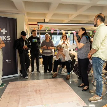Bir mekan içindesiniz; ortada büyük bir harita yer alıyor. Haritanın etrafında insanlar toplanmış, bazıları ayakta duruyor, bazıları ise oturmuş. Duvarların birinde dikkat çekici bir banner yer alırken, diğerinde şehir temalı bir logo görülmekte. Ayakta duran bir kişi dikkatlice bir şeyler anlatıyor, diğerleri ona odaklanmış durumda. Kalabalık içinde farklı kıyafetler ve stiller dikkat çekiyor; kimileri daha rahat, kimileri ise resmi görünüyor. Halka şeklinde toplanan grup, haritanın önünde etkileşimde bulunuyor; bazıları ellerinde içecekler tutarken, biri düşünceli bir şekilde oturuyor. Mekanda renkli bayraklar ve çeşitli objeler de var, ortam canlı ve samimi bir havaya sahip.