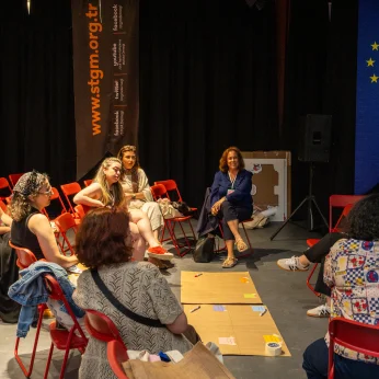 Bir grup birey, kırmızı sandalyelerle çevrili bir alanda oturmuş. Ortada, üzerinde çeşitli renklerde etiketlerin ve yazıların bulunduğu birkaç ahşap parça var. Bazı katılımcılar, odanın arka kısmında bulunan Avrupa Birliği'ne ait bir bayrağın yanında oturuyor. Farklı tarzlarda giyinmiş olan katılımcılardan bazıları, karşılıklı olarak birbirleriyle iletişim kuruyor. Odanın arka duvarında, etkinlikle ilgili bilgilerin yer aldığı bir afiş dikkat çekiyor. Genel atmosfer samimi ve etkileşim dolu; katılımcıların beyin fırtınası yaptığı veya tartışma yürüttüğü izlenimi var. Etraf karanlık ama sahne aydınlatması bölümdeki odak noktasını vurguluyor.